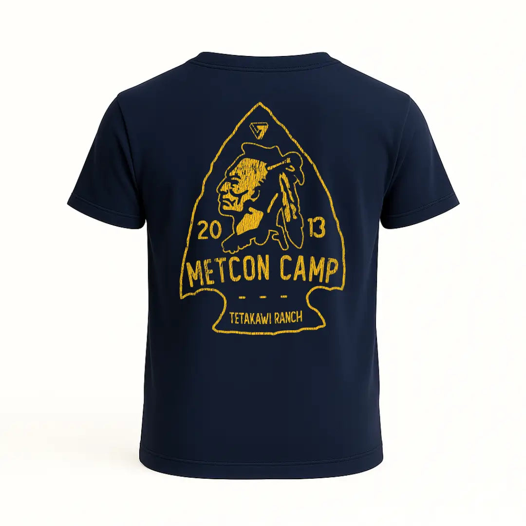 Playera azul marino con diseño trasero en forma de punta de flecha, figura de guerrero nativo y texto “Metcon Camp”, “2013” y “Tetakawi Ranch” en color amarillo. Estética rústica y tribal.