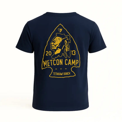 Playera azul marino con diseño trasero en forma de punta de flecha, figura de guerrero nativo y texto “Metcon Camp”, “2013” y “Tetakawi Ranch” en color amarillo. Estética rústica y tribal.