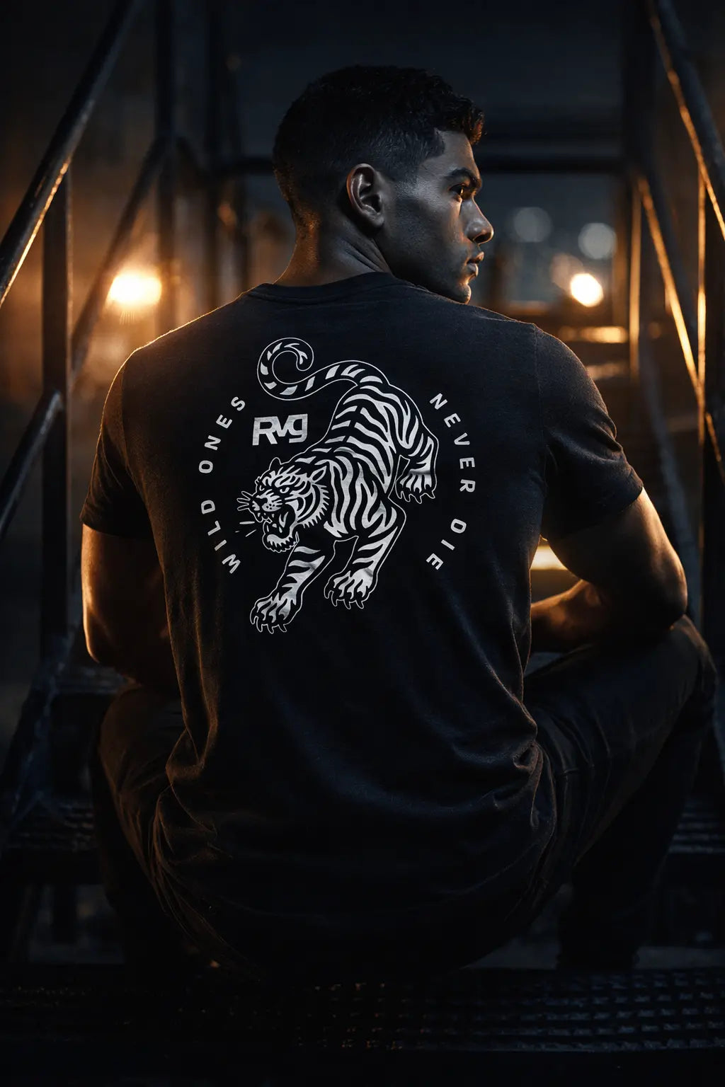 Modelo hombre con playera RVG Wild Ones RAVGE, vista trasera diseño tigre Wild Ones Never Die, ambiente industrial oscuro, ropa MMA gym